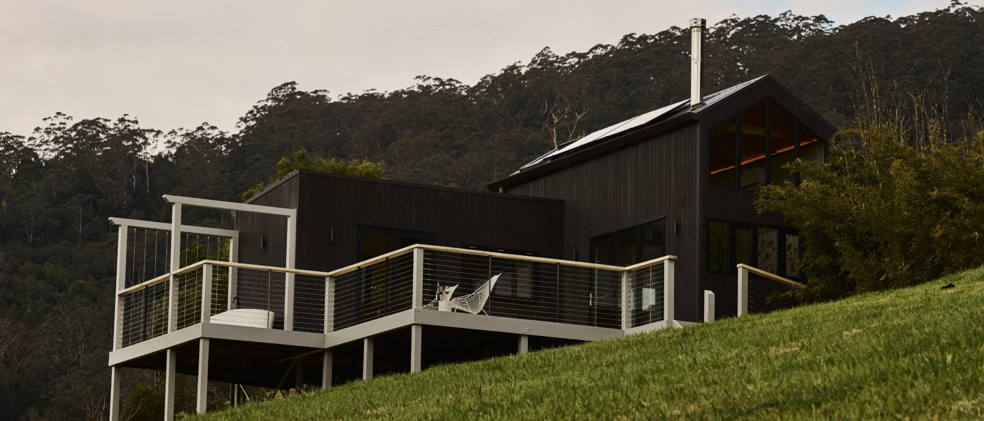 Silent Echo Cabin on its hillside in Kangaroo Valley, charcoal-black cladding above a wraparound deck, the escarpment behind